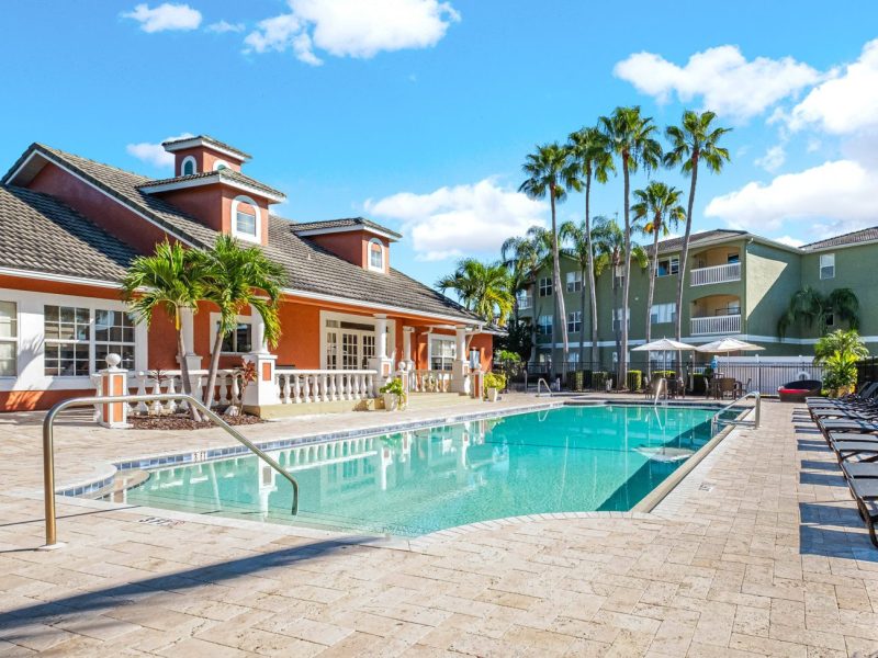 Luxury swimming pool and clubhouse at TGM University Park with palm trees and lounge chairs.