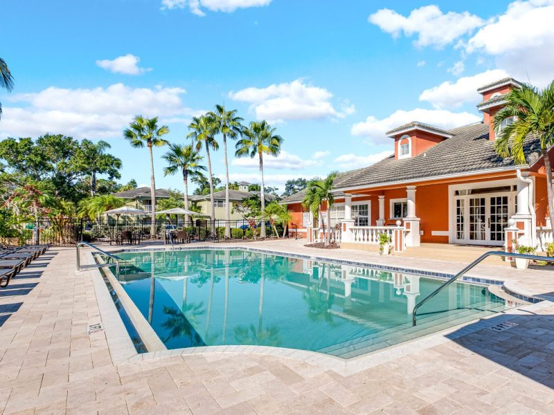Resort-style swimming pool and clubhouse at TGM University Park with palm trees and lounge chairs.